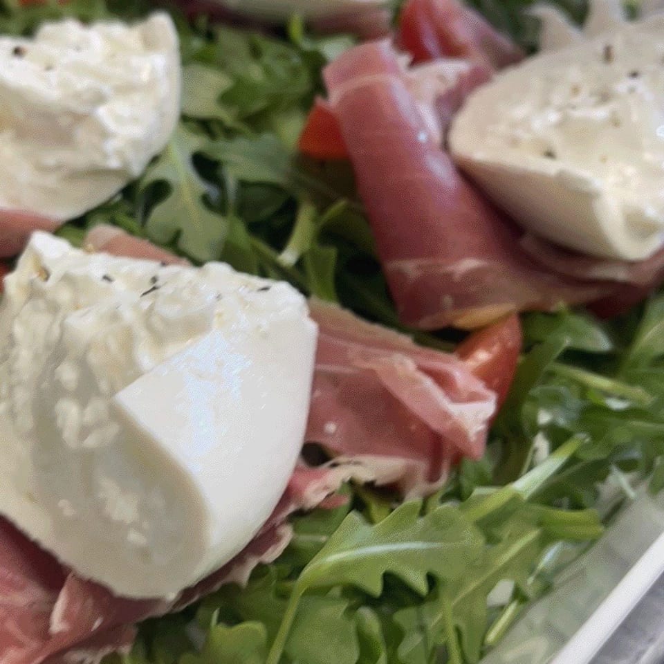 Arugula salad topped with creamy burrata cheese, prosciutto, and sliced tomatoes in a serving dish.