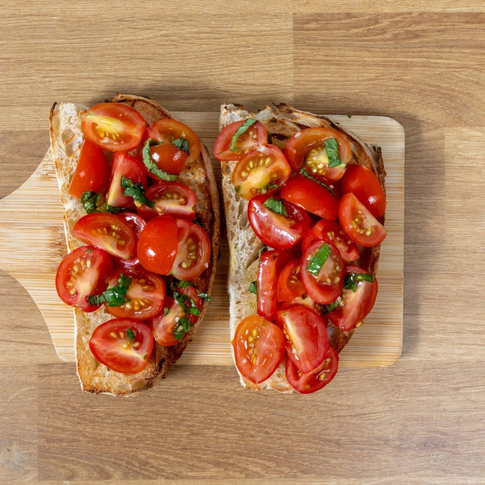Two slices of toasted bread topped with chopped tomatoes and fresh basil on a wooden cutting board.