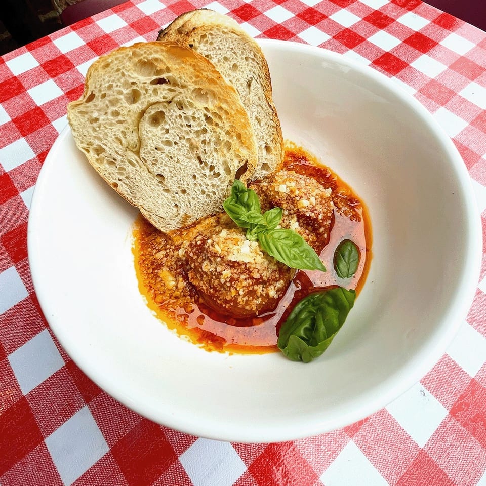 Italian meatballs topped with grated cheese and basil in tomato sauce, served with crusty bread on a red-and-white checkered tablecloth.