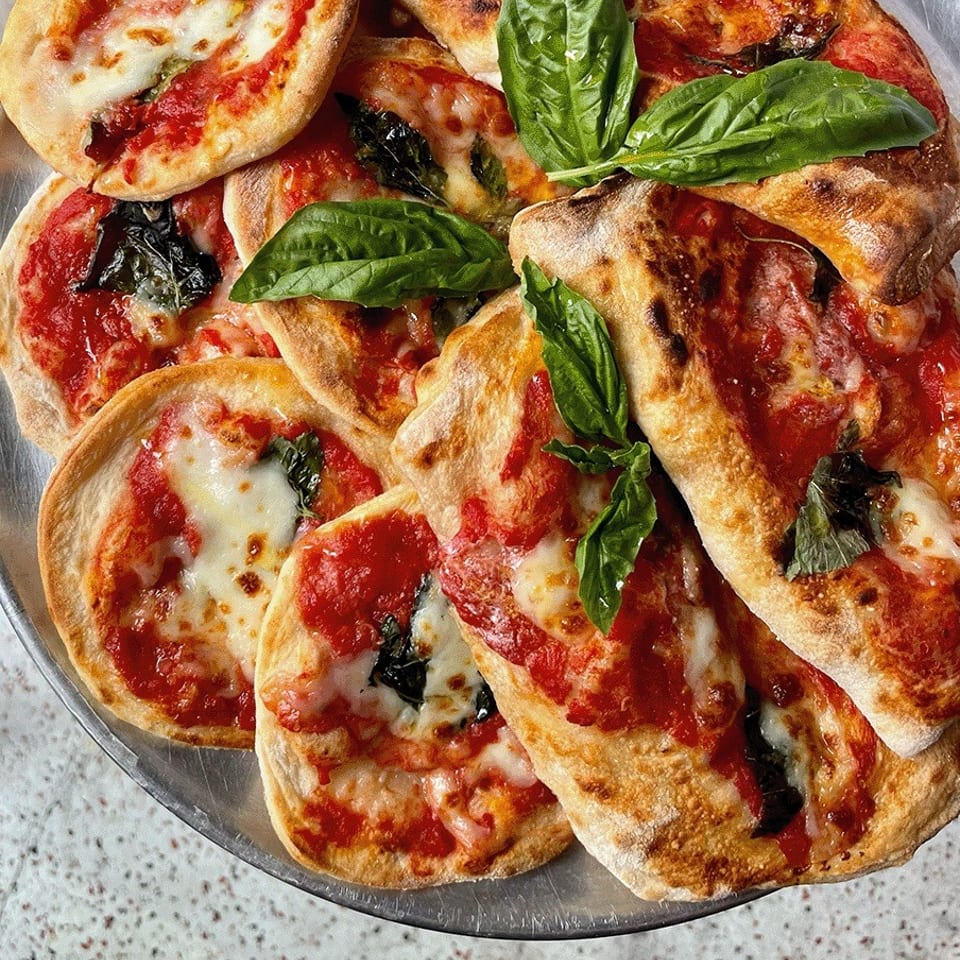 Assorted small pizzas topped with tomato sauce, melted mozzarella, basil leaves, and charred crusts on a metal platter.