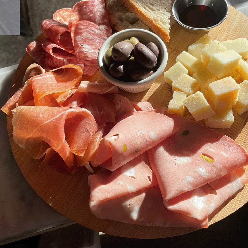 Charcuterie board with sliced cured meats, cheese cubes, olives, rustic bread, and dipping sauce in sunlight.