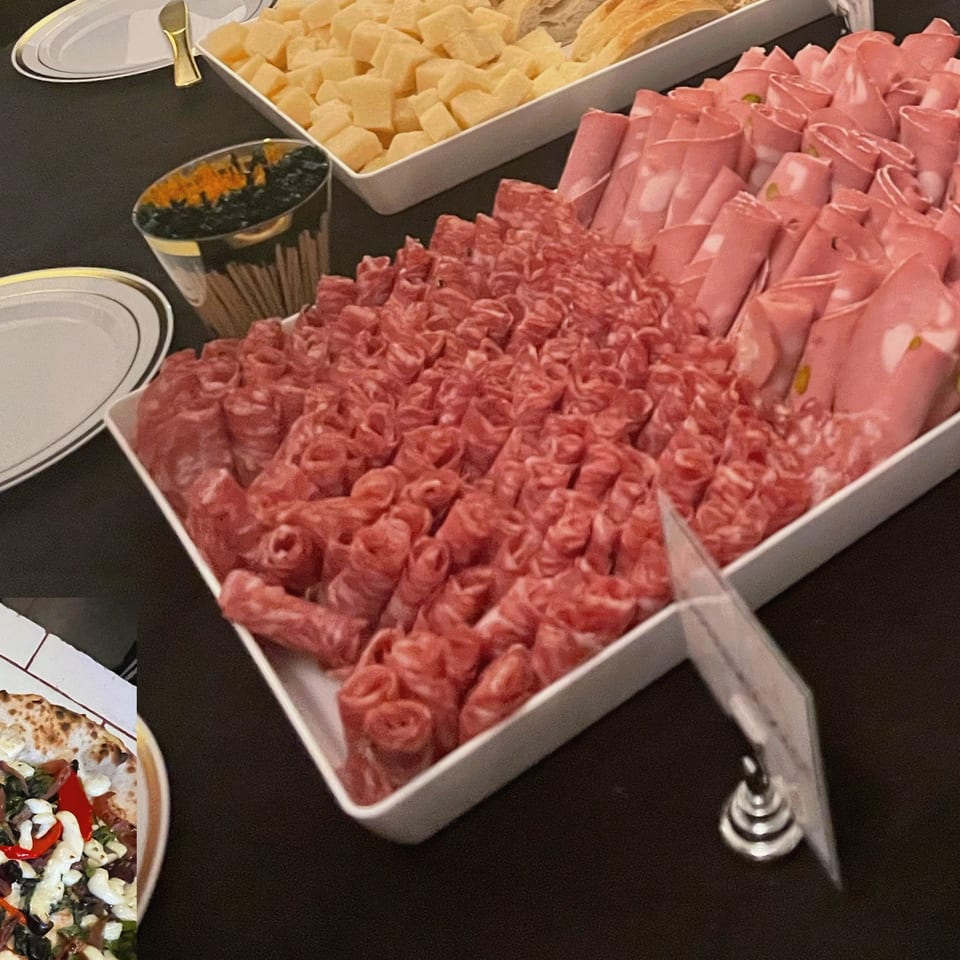 Buffet trays display rolled pink deli meats, sliced cheese, bread, crackers, and a bowl of black olives on a dark table.