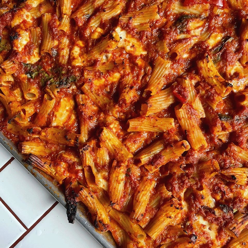 Close-up of baked rigatoni casserole with tomato sauce, melted cheese, roasted vegetables, and browned edges in a metal tray.