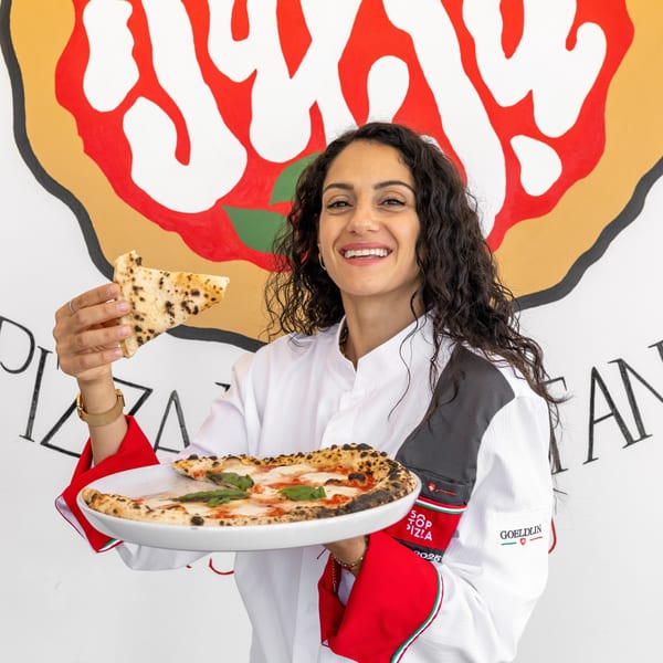 Smiling chef in white and red uniform holding a Margherita pizza and a slice.