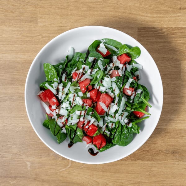 Spinach and watermelon salad with shaved cheese and balsamic glaze in a white bowl.