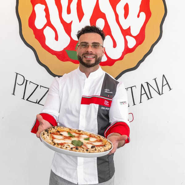 A smiling chef in a white and gray uniform holds a freshly baked Margherita pizza.