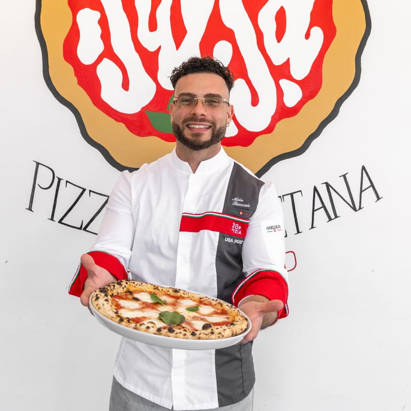A smiling chef in a white and gray uniform holds a freshly baked Margherita pizza.