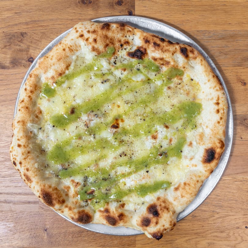 A white pizza with a green pesto drizzle and black pepper on a metal tray.