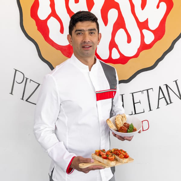 Chef holding a bowl of meatballs and a wooden board with bruschetta, against a pizza-themed wall.