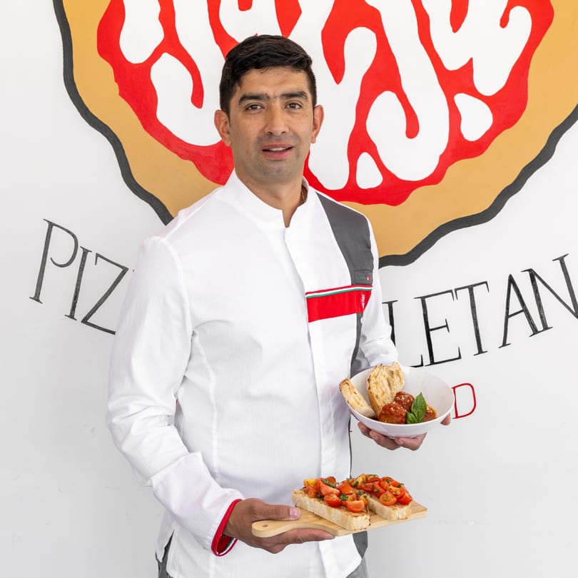 Chef holding a bowl of meatballs and a wooden board with bruschetta, against a pizza-themed wall.
