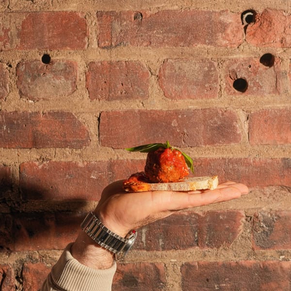 A hand holds a slice of bread topped with a meatball, sauce, and basil against a brick wall.