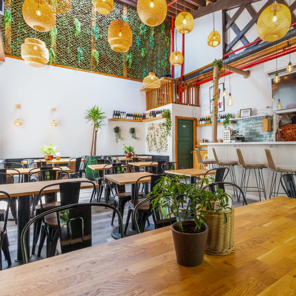 A bright, modern restaurant interior with wooden tables, black chairs, and a bar area with stools.