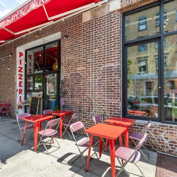 A brick pizzeria with a red awning and outdoor seating featuring red tables and purple chairs.