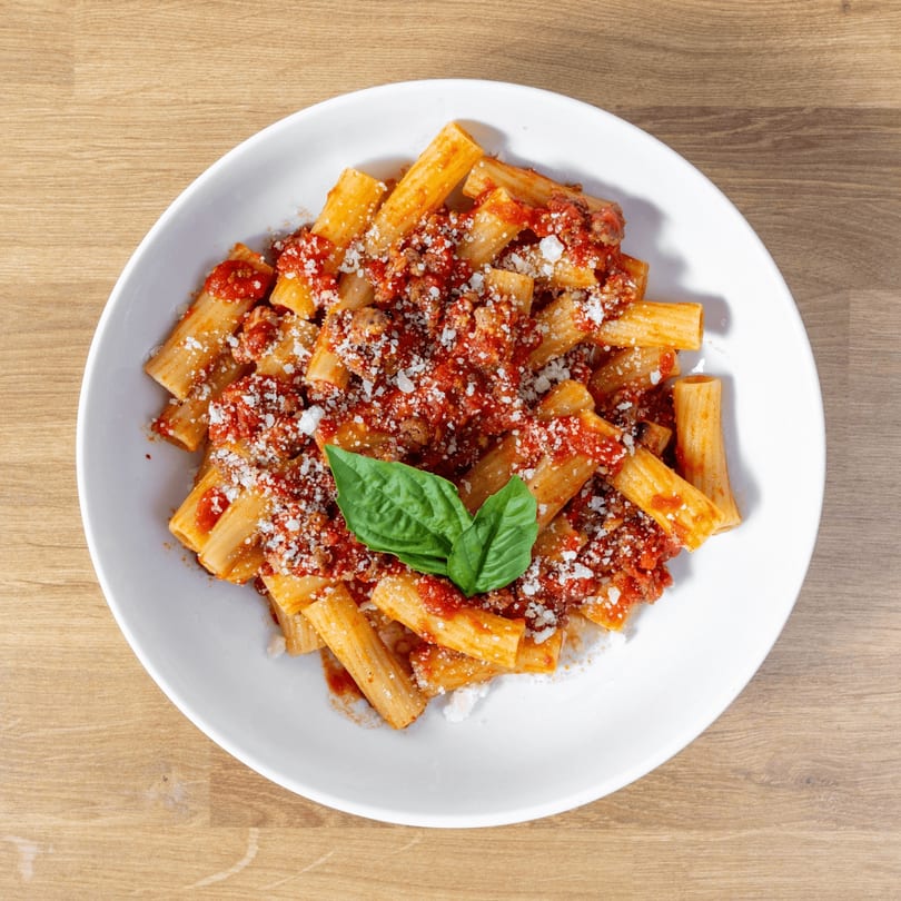 Rigatoni pasta with red sauce, ground meat, grated cheese, and fresh basil in a white bowl.