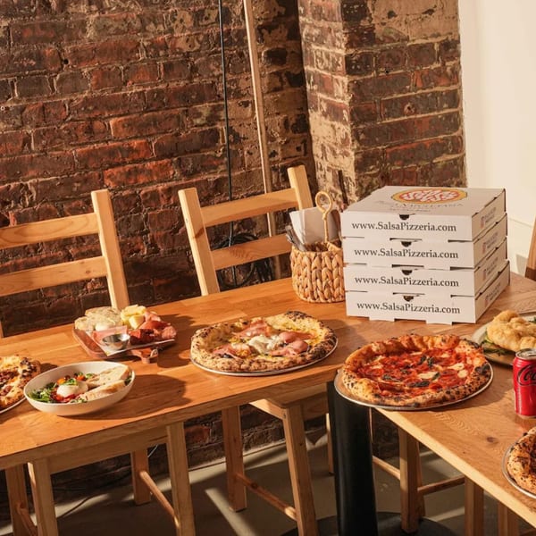 A wooden table laden with pizzas, a charcuterie board, salad, and stacked Salsa Pizzeria boxes.