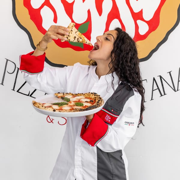 A woman in a chef's coat holds a pizza and takes a bite from a slice.