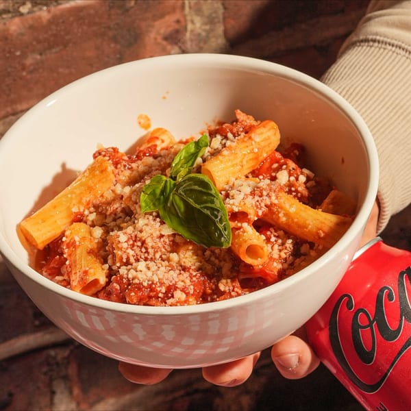A person holding a white bowl of rigatoni pasta with red sauce, parmesan, and basil, next to a Coca-Cola can.