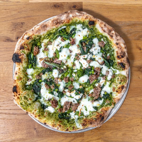 Overhead view of a pesto pizza with sausage, spinach, and ricotta cheese on a wooden table.