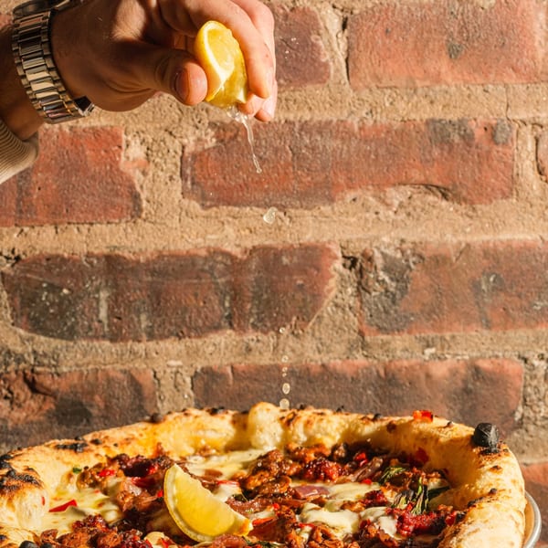 A hand squeezing a lemon over a delicious pizza with a brick wall background.