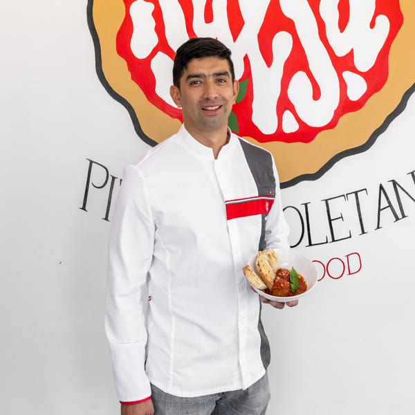 Chef in white uniform holding a bowl of meatballs with bread, against a pizza-themed wall.