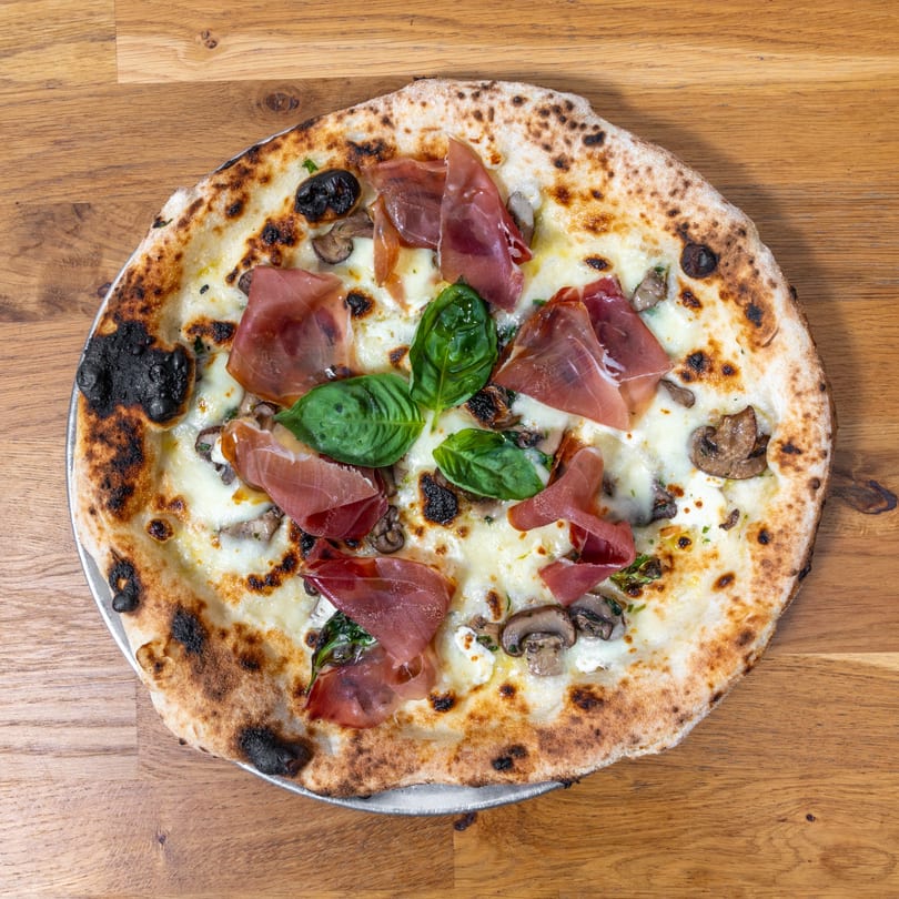 Neapolitan pizza with prosciutto, mushrooms, basil, and white cheese on a wooden table.