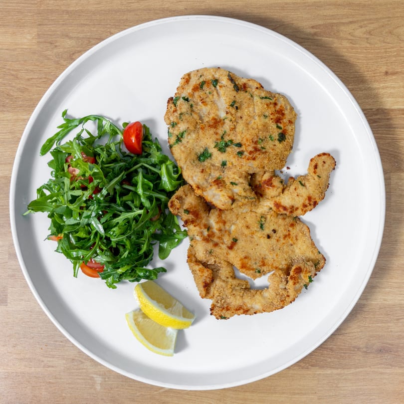 Two breaded chicken cutlets with a side of arugula salad and lemon wedges on a white plate.