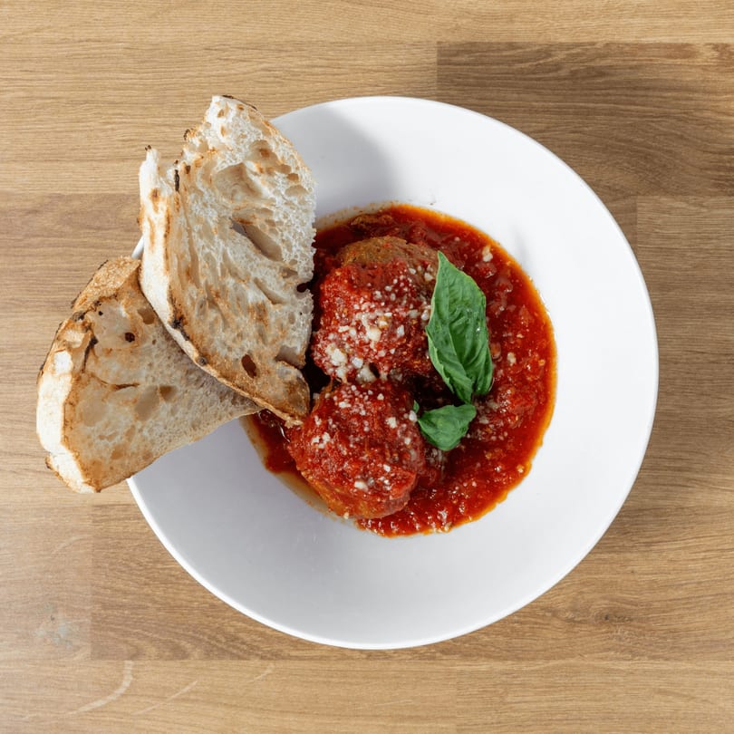 Meatballs in red sauce with grated cheese and basil, served with toasted bread on a white plate.