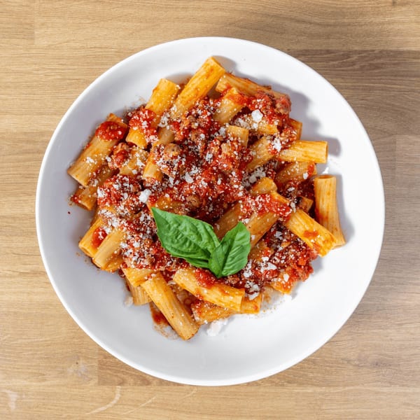 Rigatoni pasta with red sauce, ground meat, grated cheese, and basil in a white bowl.