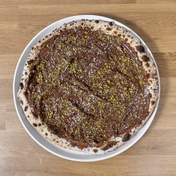 Dessert pizza with chocolate spread, powdered sugar, and pistachio crumbles on a white plate.
