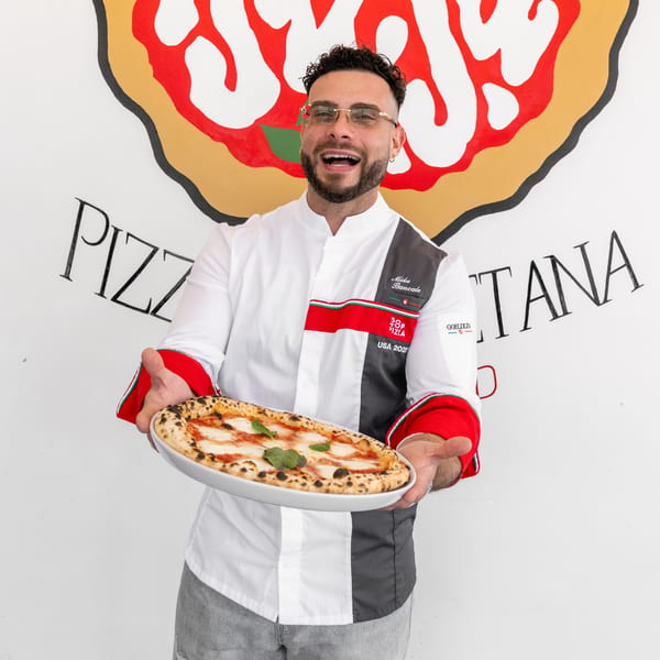 A chef in a white and gray uniform holds a freshly baked pizza with basil and mozzarella.