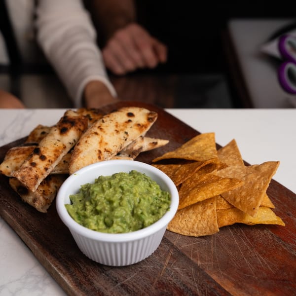 Guacamole in a white bowl with pita bread and tortilla chips on a wooden board.