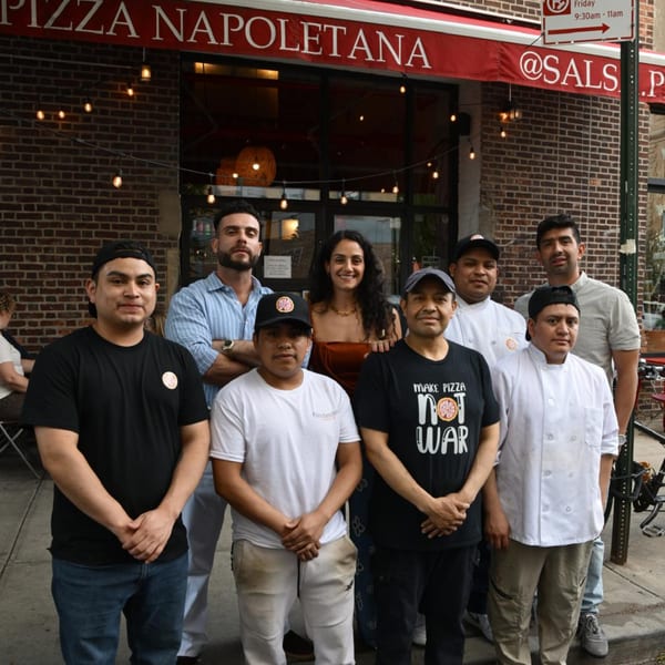 Eight restaurant staff members, including chefs, stand smiling outside a brick pizza restaurant.